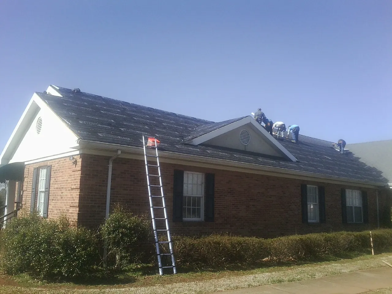 Crew tearing shingles off a brick ranch roof with a ladder set — Emergency Roof Repair work by our crew in Union Springs