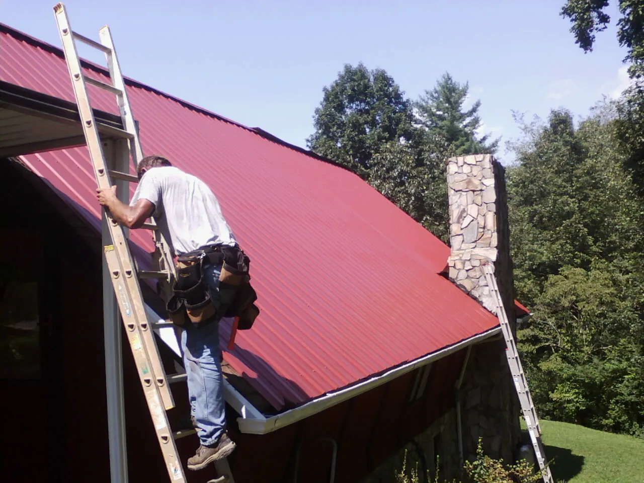Roofer on a ladder beside a red metal roof and stone chimney — Soffit & Fascia Repair by Roofing Brothers in Union Springs